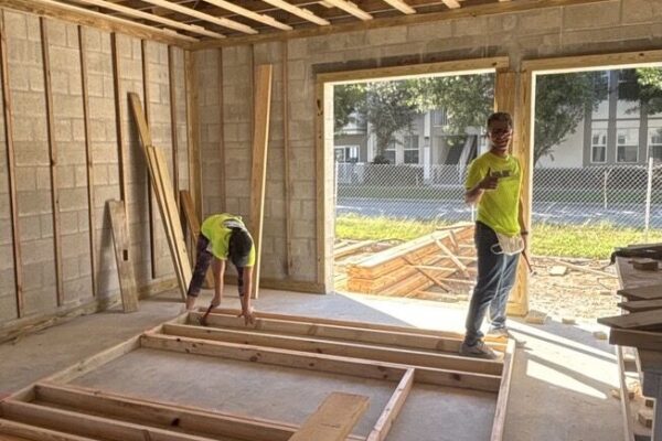 Two people in neon shirts build a wooden frame indoors.