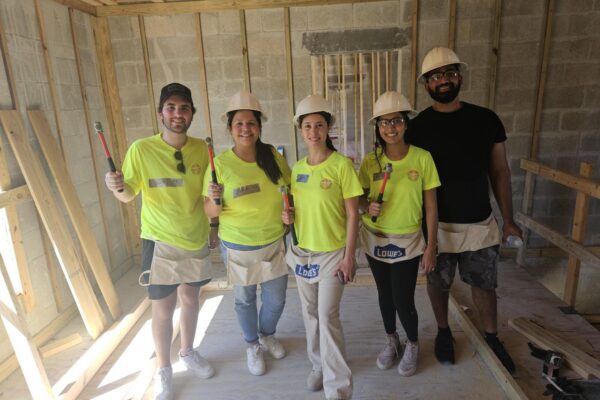 Five workers in hard hats and yellow shirts smile, holding tools.