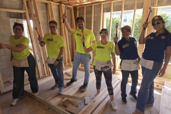 Six people with hammers pose inside a wooden building under construction.