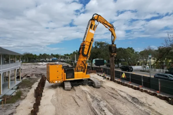 Yellow pile driver installs beams; worker in vest watches.