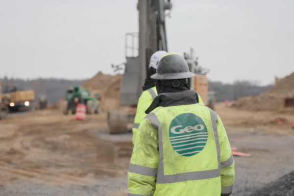 Two workers in safety gear on dirt site with machinery.