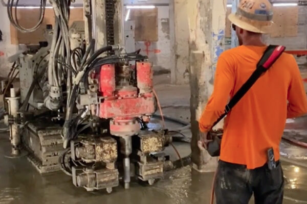 Worker in orange shirt drills on wet concrete inside building.