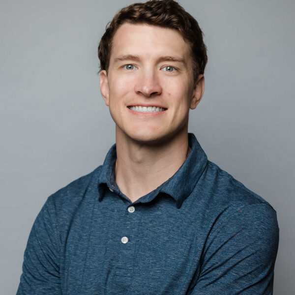 Smiling man with short brown hair in blue shirt, gray background.