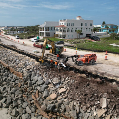 Workers repair seawall with heavy machinery near homes and rocks.