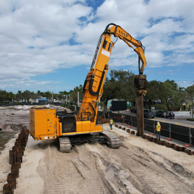 Yellow excavator installing sheet piles by road, workers nearby.