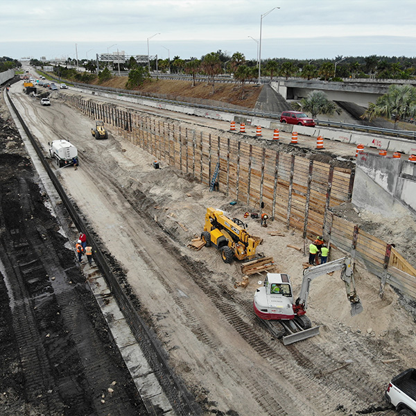 Workers and machines build a highway wall; cones and cars nearby.