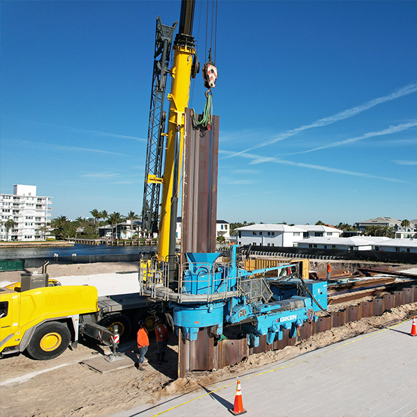 Crane lifting steel sheet pile at waterfront, workers nearby.
