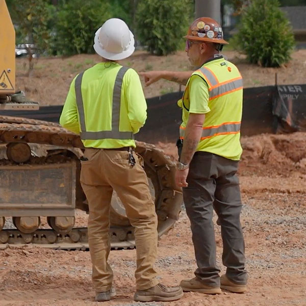 Two construction workers in safety gear talk beside equipment.