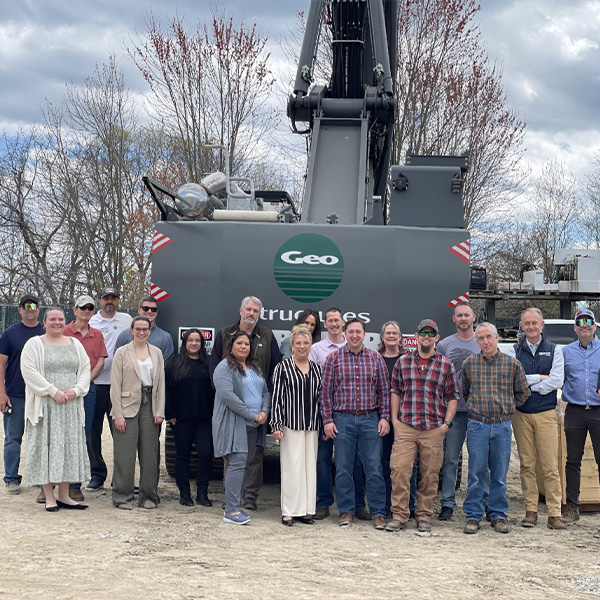 People in front of a large Geo construction vehicle outdoors.