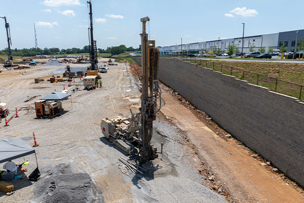 Heavy machinery drilling by a retaining wall at a construction site.