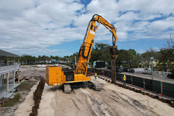 Excavator installs sheet piles at construction site by the road.