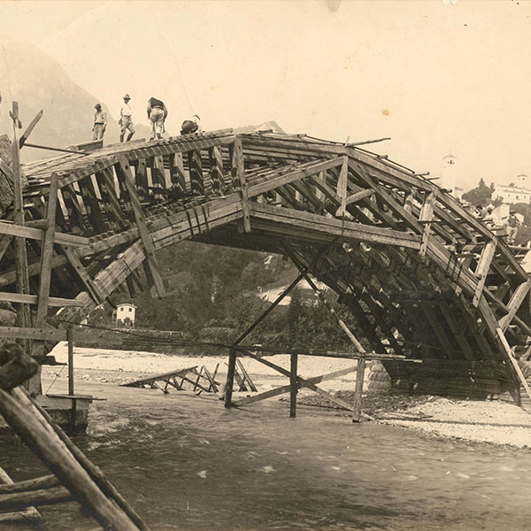 Workers build a wooden arch bridge over a river; buildings behind.