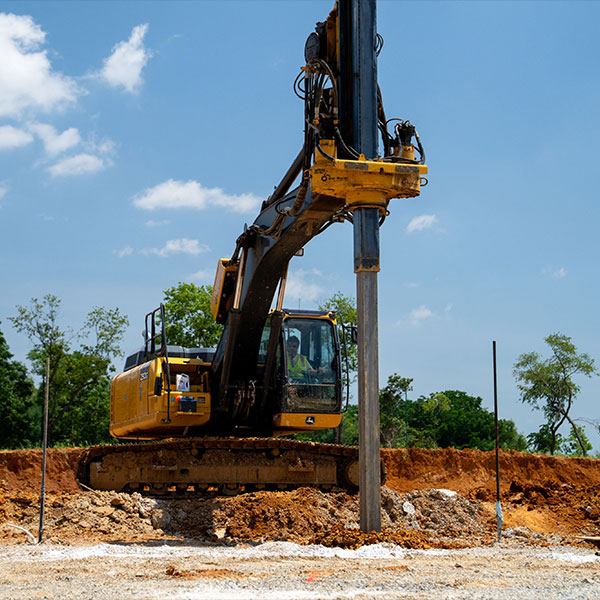 Yellow excavator driving concrete pile into ground on sunny day.