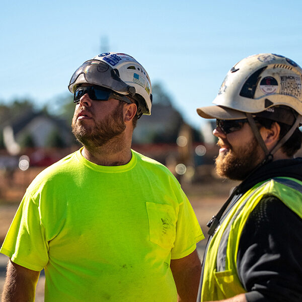 Two construction workers in safety gear look up at worksite.