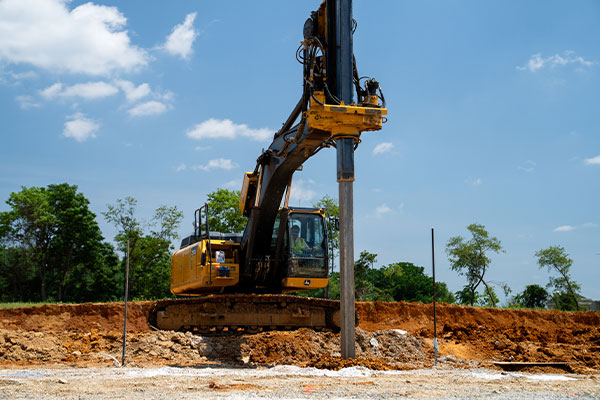 Yellow excavator drives piling into ground at construction site.