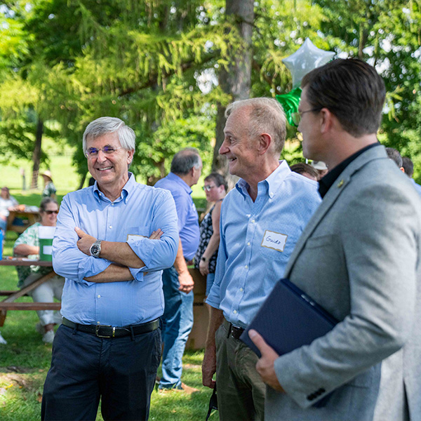 Three men talk outdoors near picnic tables and trees.