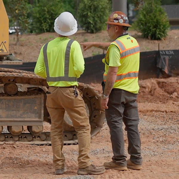 Two construction workers in safety gear stand by heavy equipment.