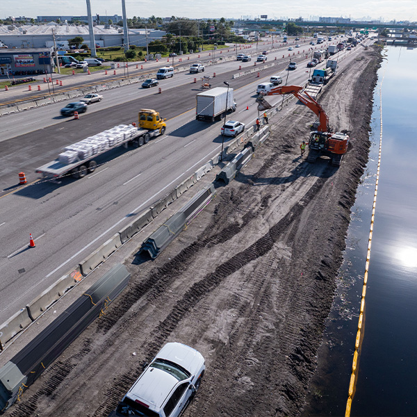 Excavator at site near highway, water, and passing vehicles.