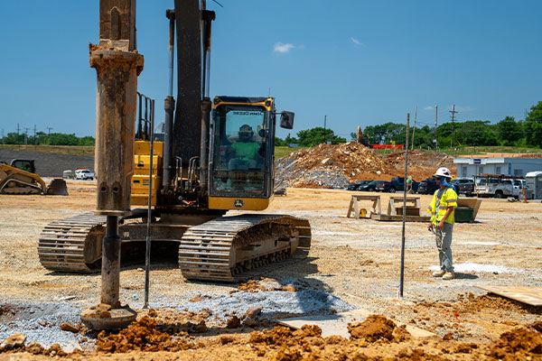 Construction worker by excavator on gravel lot, clear sky.