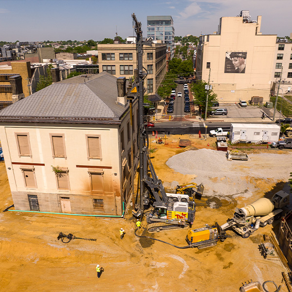 Workers and machines at construction site beside old building; vehicles nearby.