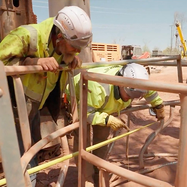 Workers in safety gear measure a bent metal railing outdoors.