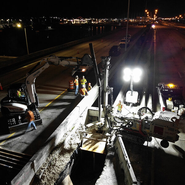 Workers use machinery on a lit highway at night.