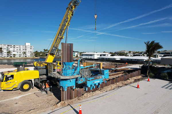 Crane installs sheet piling at waterfront; workers, cones visible.