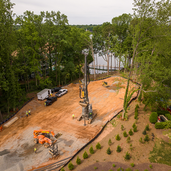 Construction site by lake with machinery, workers, and trees.