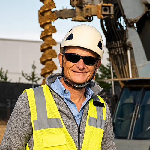 Man in a hi-vis vest with sunglasses and a white hard hat on with a construction background