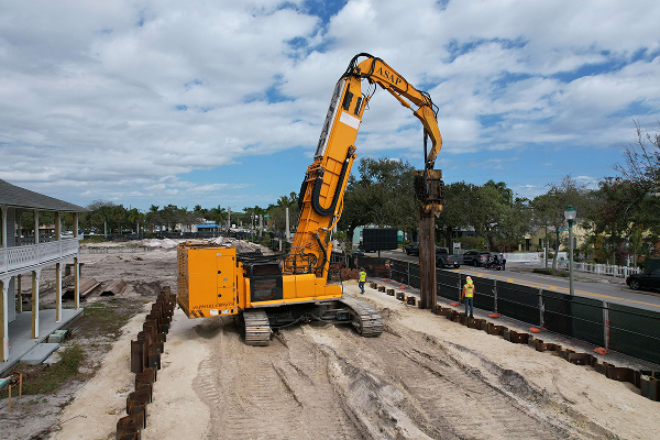 Yellow excavator installs steel piles; two workers watching nearby.