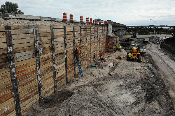 Workers reinforce wooden wall near roadwork, machinery, and barriers.